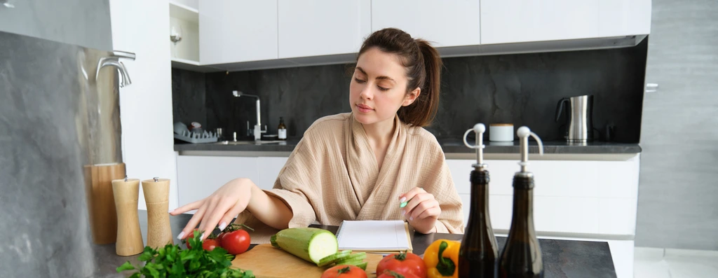 Charlotte Hayes at her kitchen counter, writing down a new recipe in her notebook with fresh ingredients in front of her.