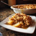 A close-up of a warm, comforting slice of apple crumble with a golden brown topping on a white plate.