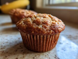 Close-up of a golden-brown banana protein muffin with a textured top, in a paper liner.