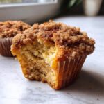 Close-up of a coffee cake muffin with a bite taken out, showing a crumbly streusel topping and moist interior.