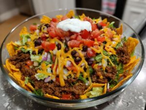 A large glass bowl filled with Dorito Taco Salad, featuring seasoned ground beef, lettuce, black beans, tomatoes, cheese, and a dollop of sour cream.