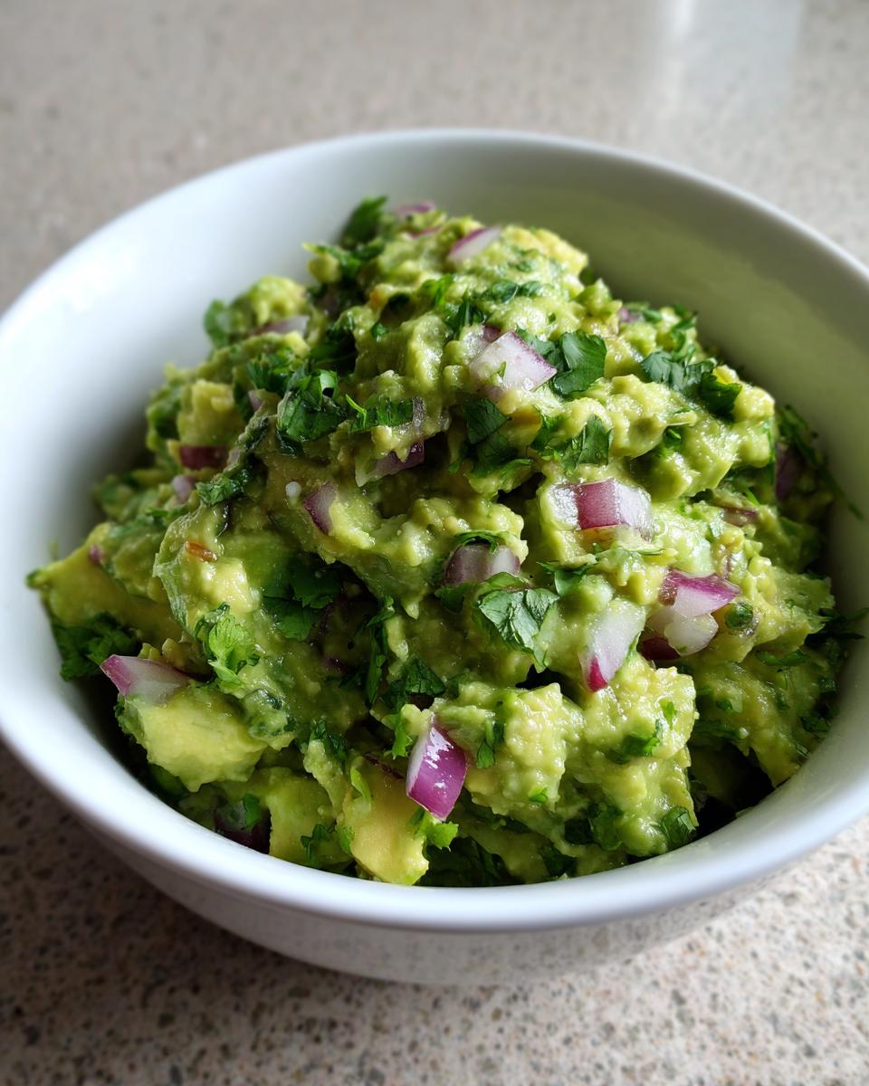 Close-up of chunky Homemade Guacamole in a white bowl, with visible avocado chunks, red onion, and cilantro.