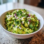 A close-up of a white bowl filled with chunky, vibrant green Homemade Guacamole, topped with chopped red onion and cilantro.