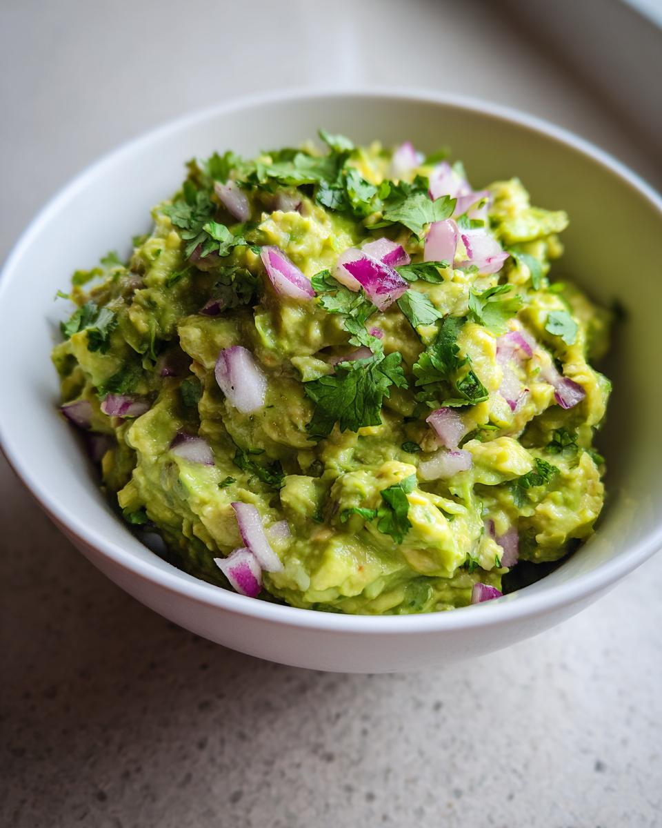 A close-up of a bowl of vibrant green Homemade Guacamole, topped with diced red onion and fresh cilantro.