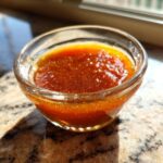 A small glass bowl filled with glistening honey BBQ sauce, showcasing its rich amber color and slightly textured surface.