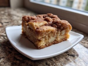 A square slice of moist apple snickerdoodle bars topped with cinnamon sugar, served on a small white plate.