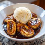 Close-up of caramelized bananas foster slices swimming in dark sauce, topped with a scoop of vanilla ice cream.