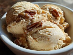 Close-up of creamy butter pecan ice cream scoops in a white bowl, topped with pecans and caramel sauce.