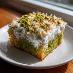A square slice of coconut and pistachio pudding cake topped with white frosting, chopped pistachios, and toasted coconut flakes.