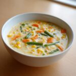 A close-up of a creamy vegetable soup filled with bright orange carrots, green beans, corn, and peas.
