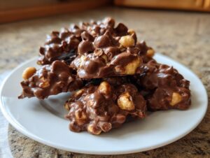 A mound of homemade Crockpot candy clusters made with chocolate and peanuts piled on a white plate.