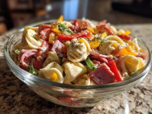 A close-up of a glass bowl filled with Italian grinder tortellini salad, featuring tortellini pasta, colorful bell peppers, and deli meats.