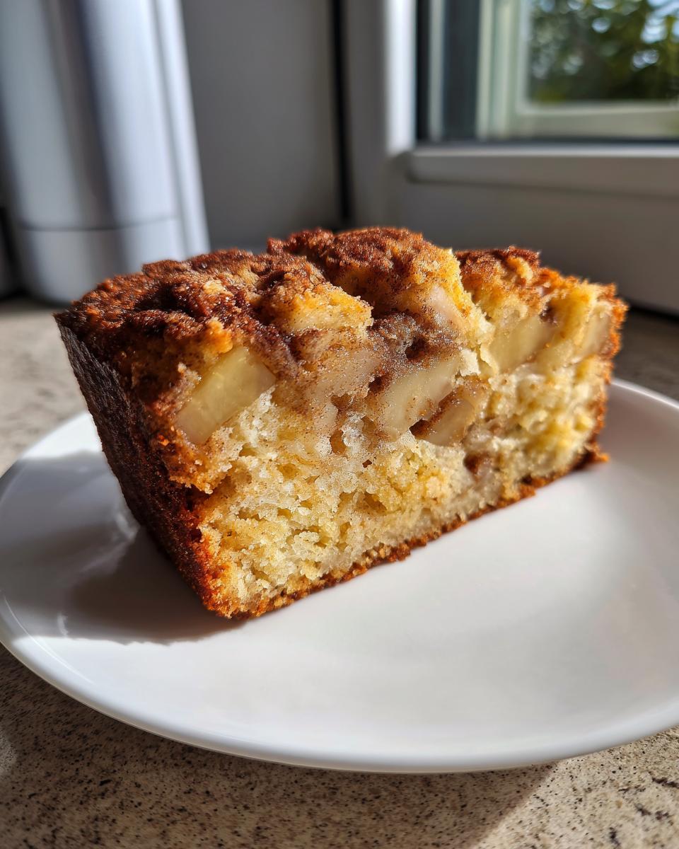 A close-up of a moist slice of apple cake with visible apple chunks and a cinnamon topping on a white plate.