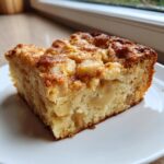 A close-up of a moist slice of apple cake, featuring visible chunks of apple and a cinnamon crumble topping.