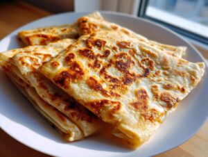 A stack of golden, flaky Moroccan chicken msmen squares with browned spots, resting on a white plate.