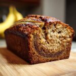 A close-up of a freshly baked protein banana bread loaf on a wooden cutting board, with bananas in the background.