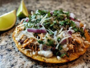 A close-up of a delicious rotisserie chicken tostada topped with cheese, red onion, and cilantro, with lime wedges on the side.