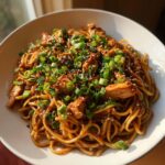A bowl filled with saucy sticky garlic chicken noodles, topped generously with sliced green onions and sesame seeds.