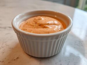 Close-up of creamy, orange-pink homemade burger sauce speckled with herbs in a small white ramekin.