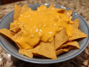 A close-up view of tortilla chips generously covered in smooth, bright orange nacho cheese sauce in a blue bowl.