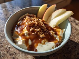 A bowl of creamy apple dip topped with caramel sauce and crunchy bits, served with apple slices.