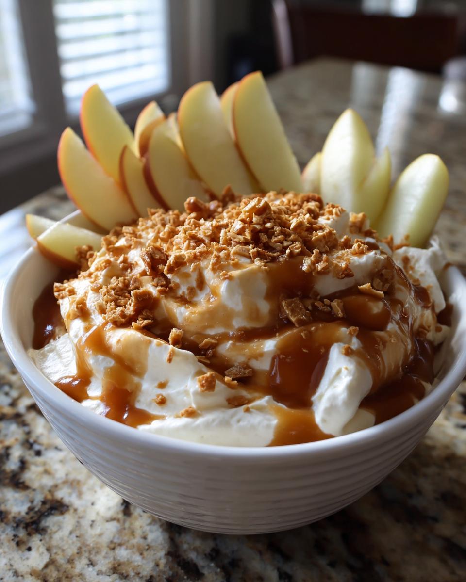A white bowl filled with creamy apple dip, topped with caramel sauce and crunchy bits, served with fresh apple slices.
