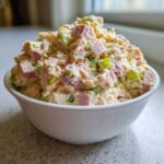 A close-up of creamy ham salad with visible chunks of ham and green celery in a small white bowl.