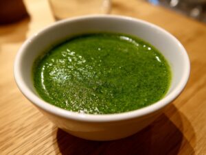 A close-up shot of vibrant green, creamy spinach soup served in a small white bowl on a wooden surface.