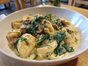 A close-up of a white bowl filled with creamy tortellini soup, featuring cheese tortellini, wilted spinach, and mushrooms.