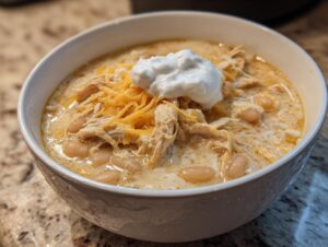 A close-up of a creamy white chicken chili bowl topped with shredded cheese, sour cream, and white beans.