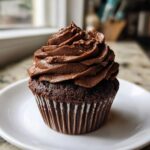 A single chocolate cupcake topped with a generous swirl of rich chocolate frosting, sitting on a white plate.