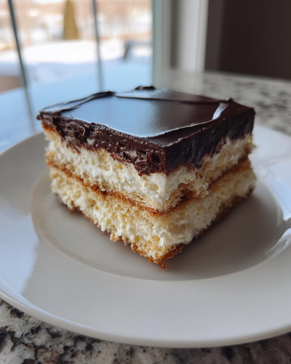A square slice of eclair cake showing layers of cake, cream filling, and a glossy chocolate topping on a white plate.