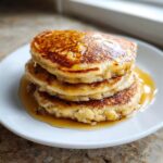 A tall stack of three fluffy banana pancakes generously drizzled with golden syrup on a white plate.