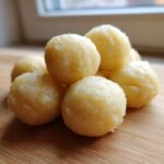 A close-up of several freshly made, round potato dumplings piled on a light wooden cutting board.