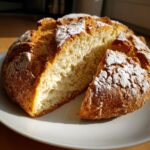 A rustic, round loaf of homemade soda bread, dusted with flour, with one slice cut and resting beside it on a white plate.