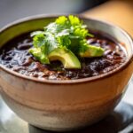Close-up of a bowl of rich, dark black bean soup garnished with fresh avocado slices and cilantro.