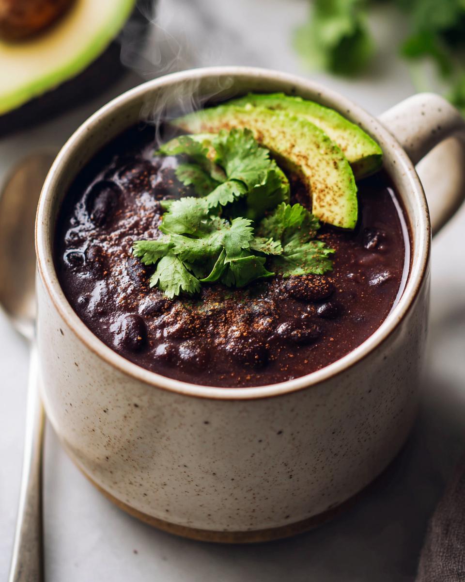 Close-up of a steaming mug filled with thick, dark black bean soup, topped with avocado slices and fresh cilantro.