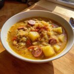 Close-up of a white bowl filled with rich, golden sauerkraut soup featuring chunks of potato, sliced sausage, and topped with paprika.