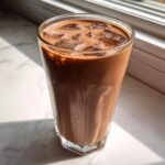 A tall glass filled with rich, iced chocolate milk, glistening in bright sunlight.
