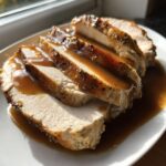 Close-up of thick slices of moist crockpot turkey breast covered in rich brown gravy on a white plate.