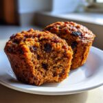 Two moist bran muffins on a white plate, one cut in half showing the texture and raisins.