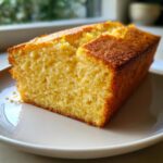 A close-up of a moist, golden slice of olive oil cake on a light gray plate near a window.