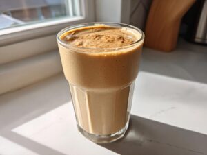 A tall glass filled with a thick, creamy peanut butter banana smoothie, dusted with cinnamon, sitting on a white counter near a window.