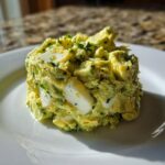 A mound of creamy, green avocado egg salad featuring visible chunks of hard-boiled egg whites, served on a white plate.