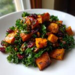 A close-up of a vibrant sweet potato salad featuring roasted orange sweet potato cubes mixed with dark green kale, dried cranberries, and nuts.