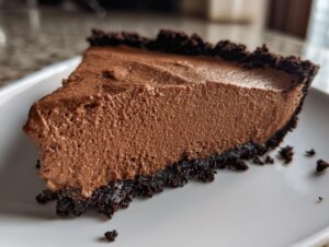A close-up shot of a thick slice of chocolate mousse pie with a dark, crumbly crust on a white plate.