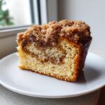 A close-up slice of moist sour cream coffee cake featuring a thick cinnamon streusel topping on a white plate.