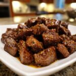 Close-up of tender, glazed cubes of crockpot steak piled on a white square plate.