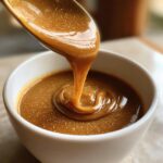 Close-up of thick, rich turkey gravy being poured from a golden spoon into a white bowl.