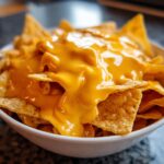 A close-up of tortilla chips completely smothered in thick, bright orange nacho cheese sauce in a white bowl.